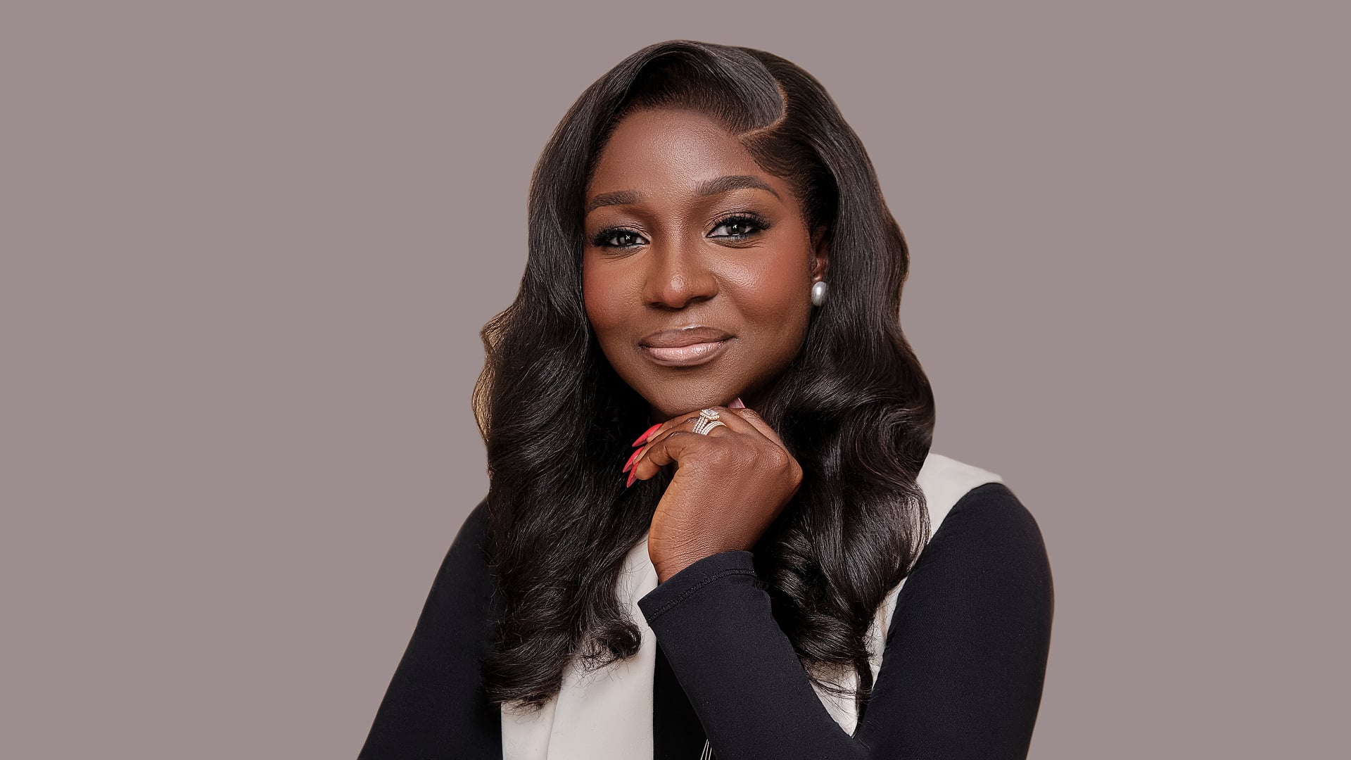 La'blinks model in a black top and cream blazer against a neutral gray background, hand resting under her chin, voluminous luxury hair